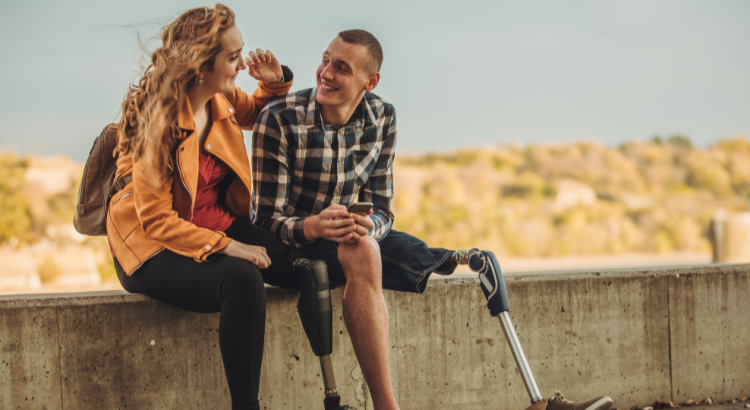 A man and a woman sitting on a low wall talking happily with sand dunes in the background. Both the man and the woman have prosthetic left legs.
