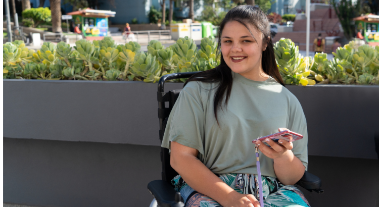 A young woman in a powered wheelchair sits in front of a grey wall with plants on top. She is smiling towards the camera, and is holding a mobile phone in her left hand