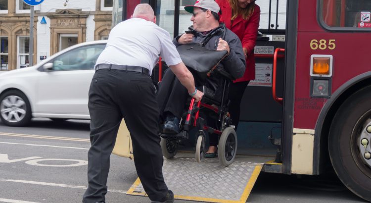 A bus driver is holding the wheelchair of a disabled person as it is cautiously lowered down a short ramp from a bus. A woman is also holding the wheelchair at the rear. The wheelchair user looks cautious. Image copyright Alamy