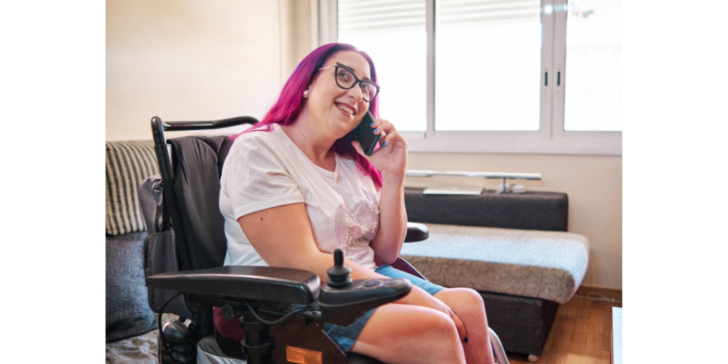 A lady with pink hair and wearing a white tshirt sits in a powered wheelchair, talking on the phone. The phone is held to her left ear, and she is smiling. There is a window and sofa in the background.
