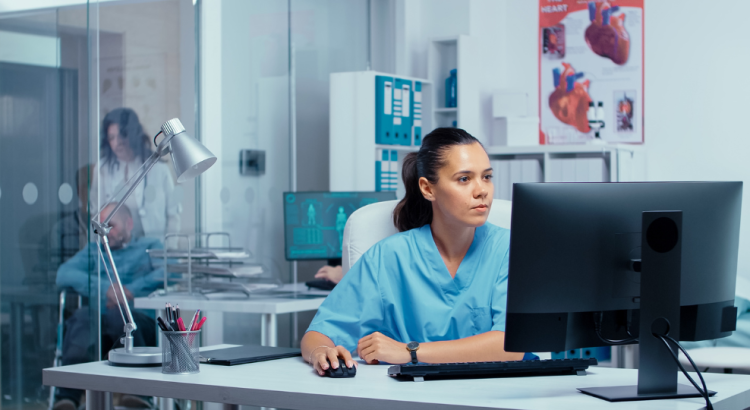 A nurse wearing blue scrubs looking intently into a large black computer monitor. In the background are medical posters and a man in a wheelchair being pushed by a female nurse.