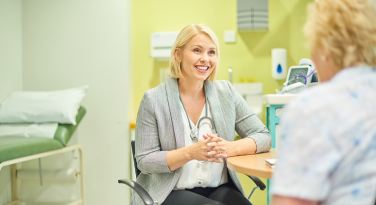 A woman wearing a white shirt and grey jacket talks to a woman wearing white, who's back is facing the camera. In the background is a medical bed set against a light yellow wall.