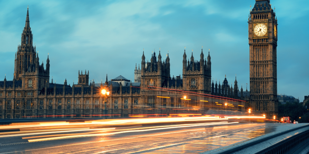 The Palace of Westminster seen from Westminster Bridge against a blue evening sky. In the foreground are coloured motion blurs of moving traffic