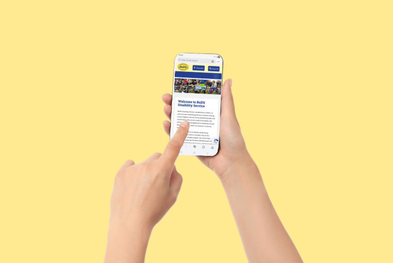 A hand holding a mobile phone showing the BuDS website homepage. The other hand has an outstretched index finger touching the phone screen.