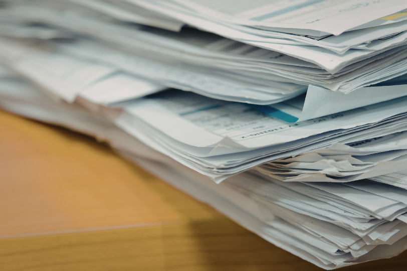 A pile of papers on a light brown wooden table. The papers are irregularly overlapping each other, and the visible faces appear to be forms.