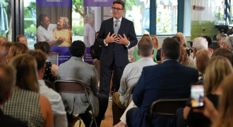 The Prime Minister, Andy Burnham, making a speech. He is a white man with dark hair and glassess dresse in a dark suit. He has his hands spread on his chest. He is standing in a care home with rows of seated people in front of him. Image copyright: BBC